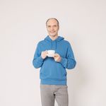 Senior man wearing a blue hoodie, holding a cup indoors, smiling casually.